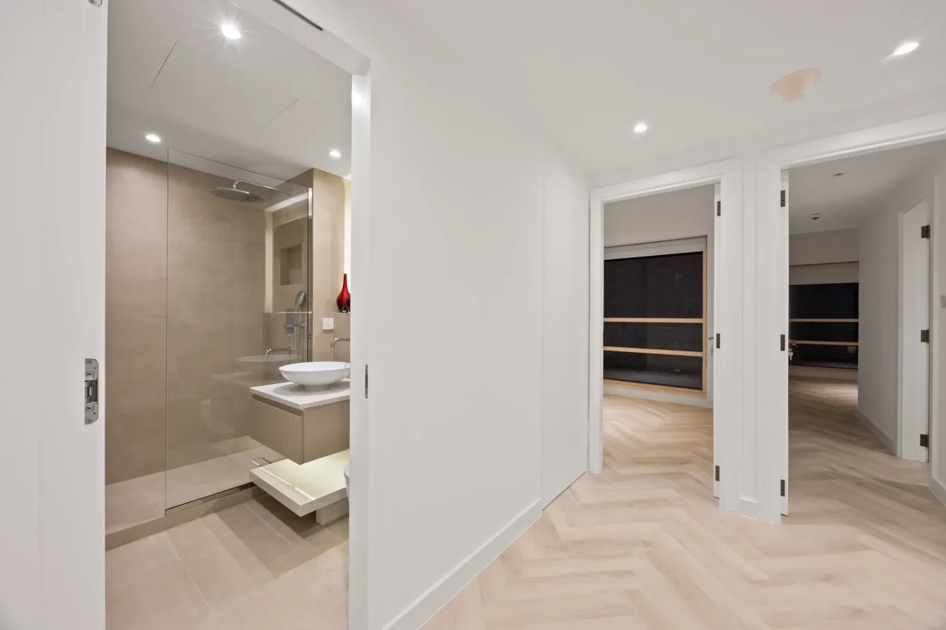 Modern hallway with open door to a beige tiled bathroom featuring a glass shower, white vessel sink, and floating vanity; white walls and warm wood floors