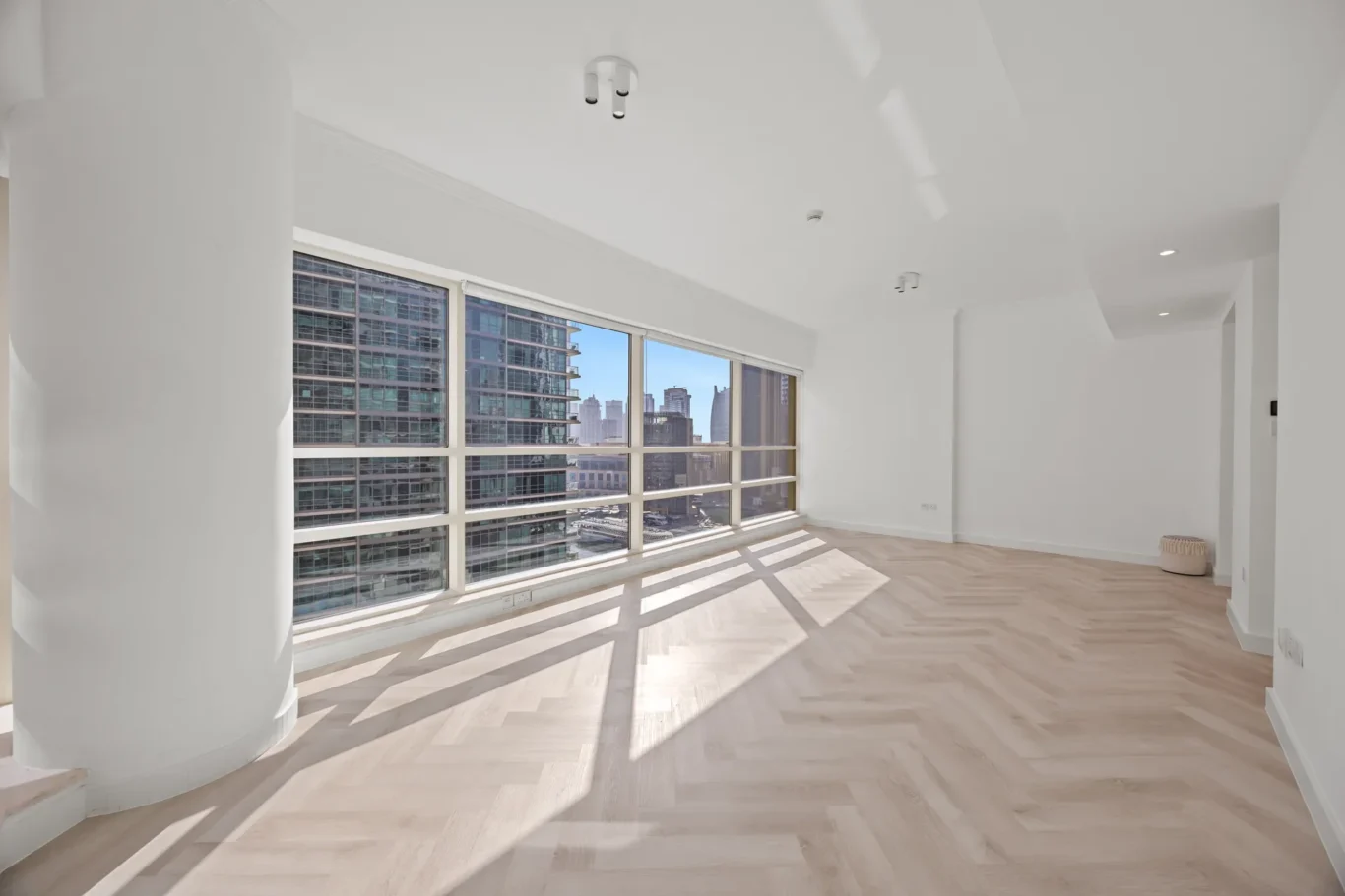 Bright empty living space with large floor-to-ceiling windows offering a city skyline view and light wood floors.