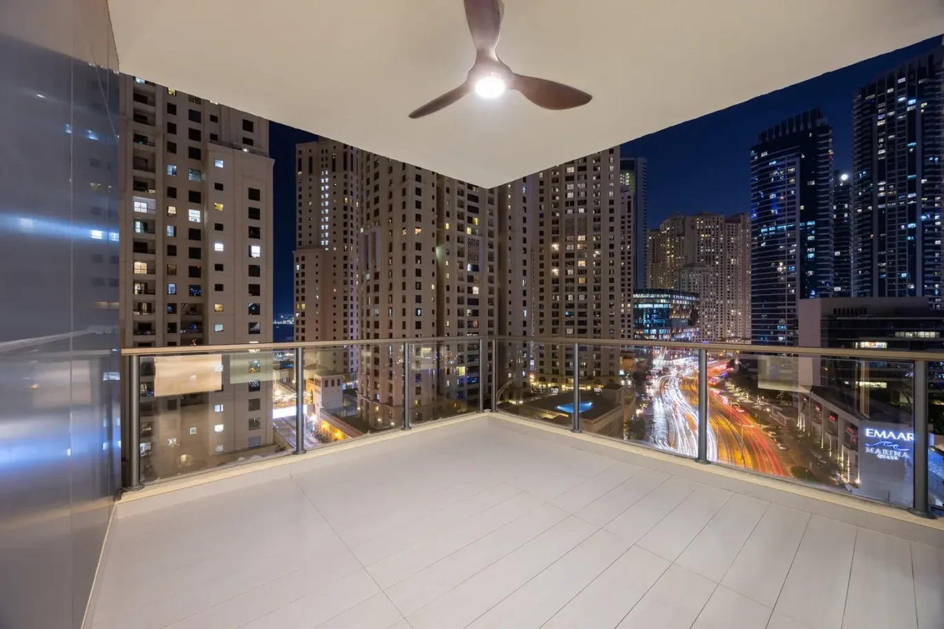 Nighttime cityscape viewed from a balcony with a glass railing and a ceiling fan overhead.