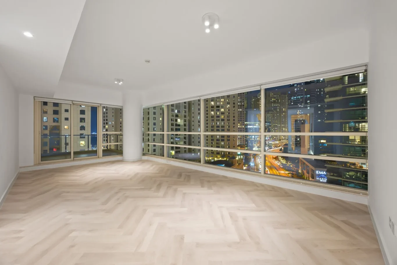 Empty modern living room at night with large floor-to-ceiling windows overlooking a city skyline, minimal decor, light wood floors.