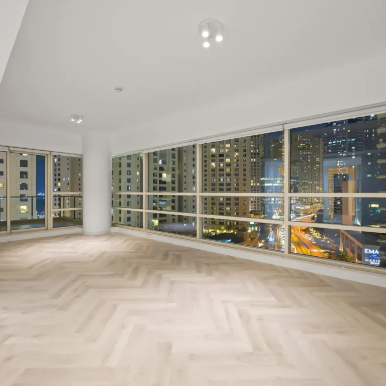 Empty modern living room at night with large floor-to-ceiling windows overlooking a city skyline, minimal decor, light wood floors.