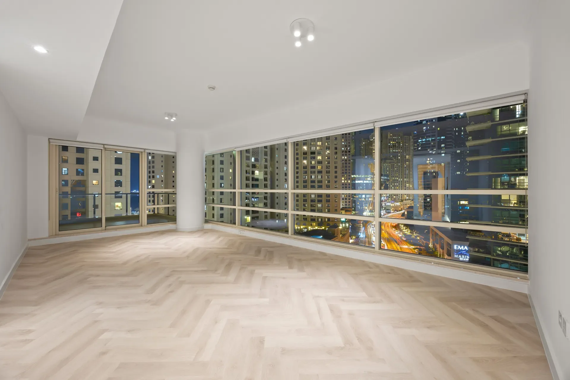 Empty modern living room at night with large floor-to-ceiling windows overlooking a city skyline, minimal decor, light wood floors.