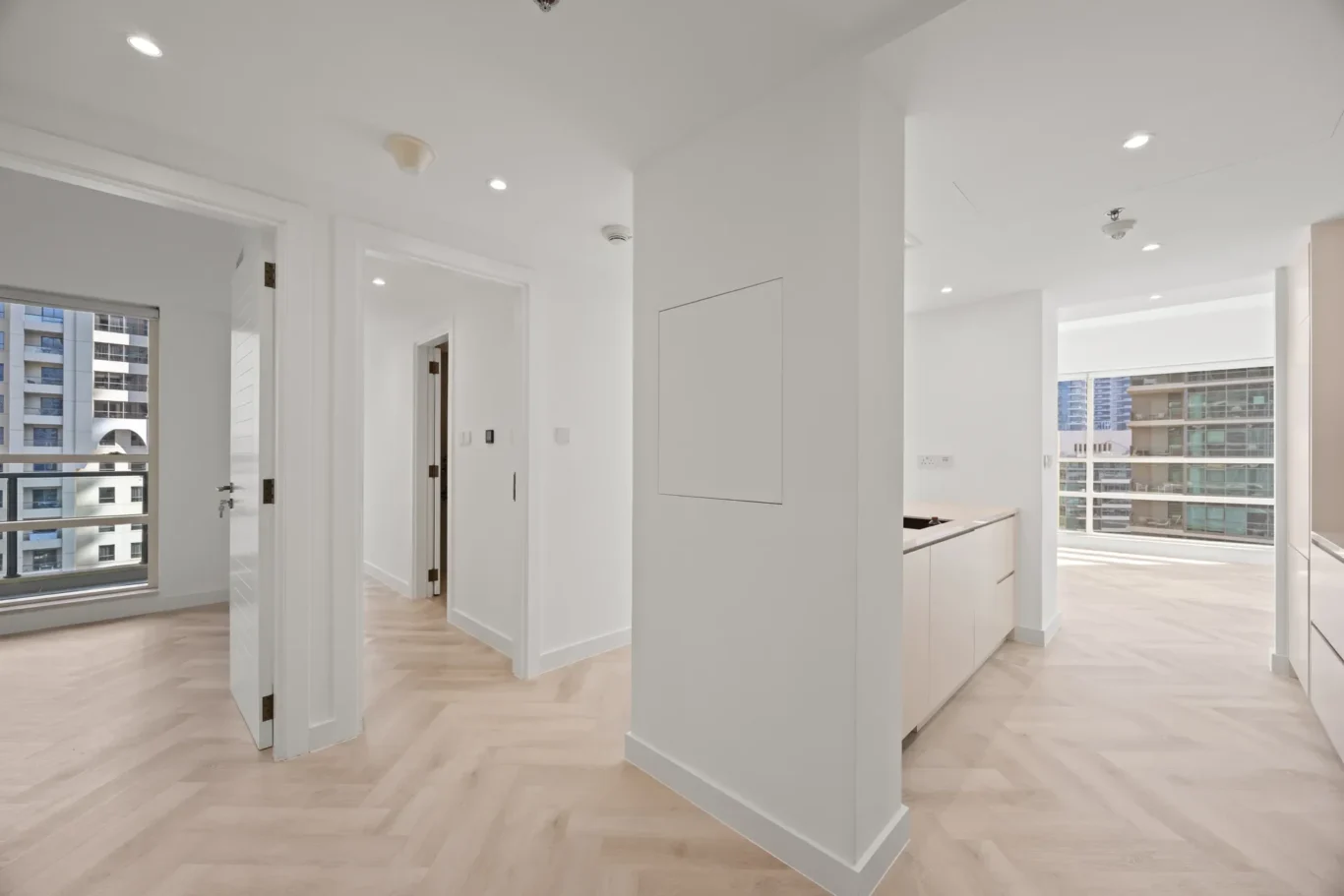 Bright, minimalist apartment interior with white walls, light wood floors, and large windows showing city buildings outside.