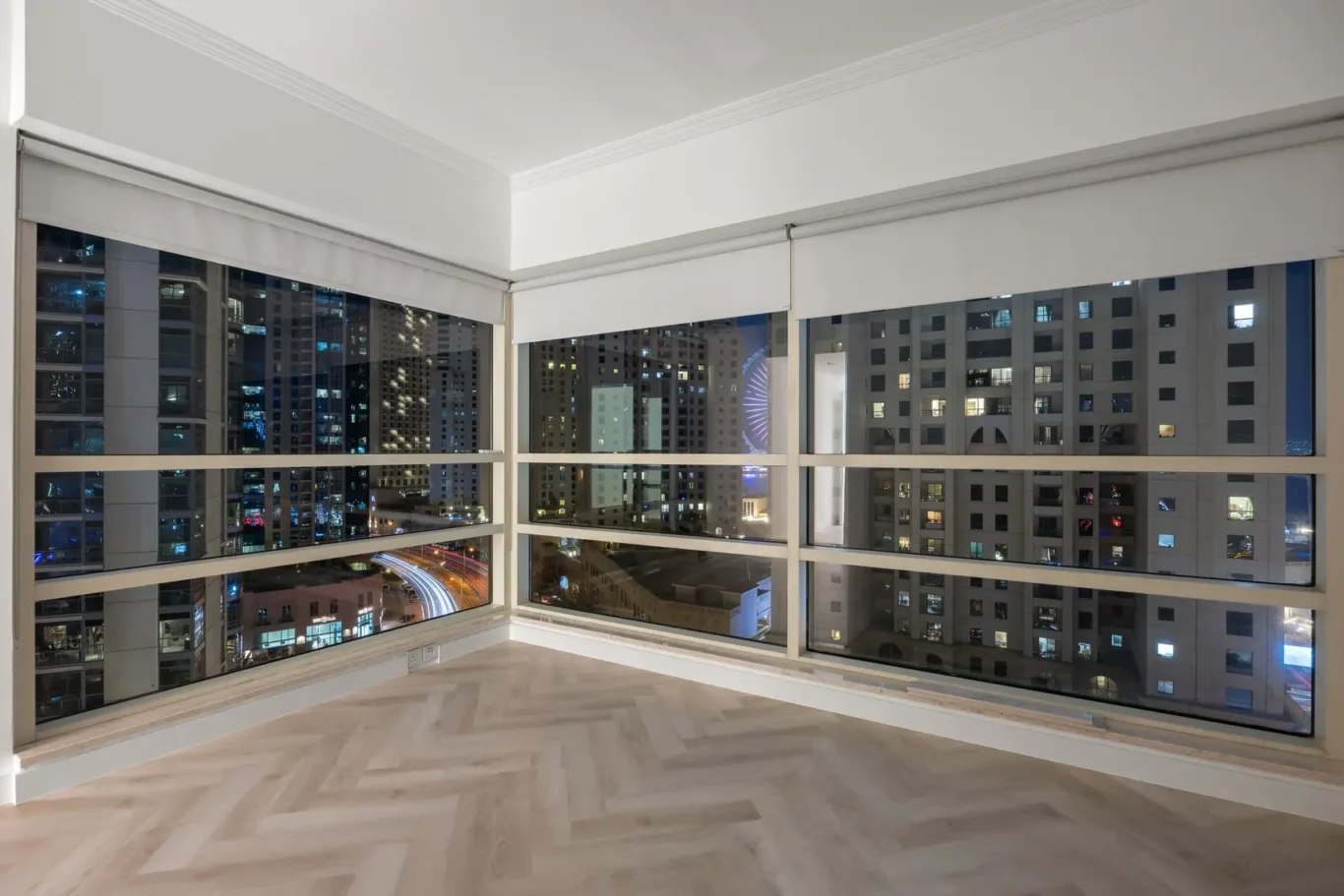 Corner room with floor-to-ceiling windows showing a nighttime cityscape and light wood herringbone floors.