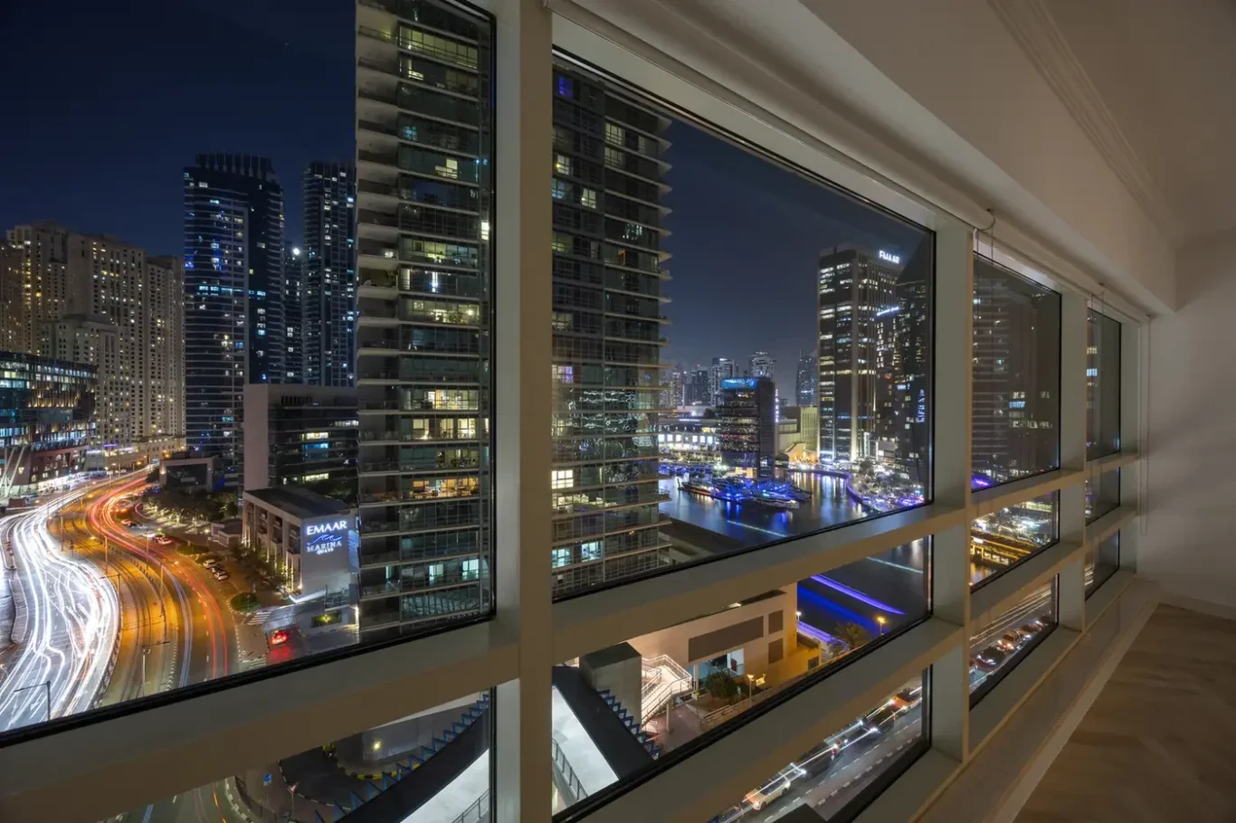 Night city skyline seen through tall window panes from a modern interior, with lit skyscrapers and a marina below, traffic light trails visible on the street edge.