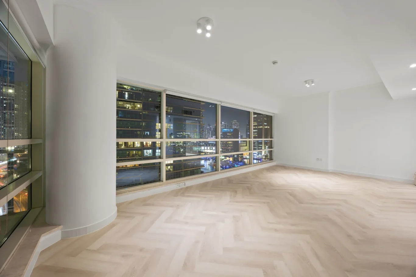Empty, modern living room with large city-view windows at night, light wood flooring and white walls (no furniture).