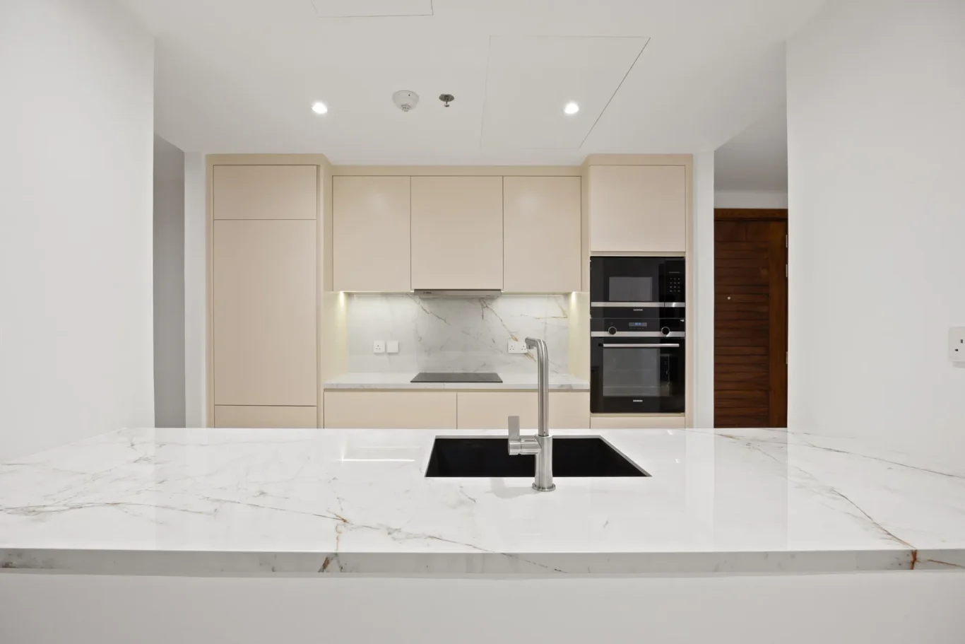 Contemporary kitchen with a marble island, beige cabinets, and built-in double ovens; a black undermount sink and chrome faucet in the foreground.