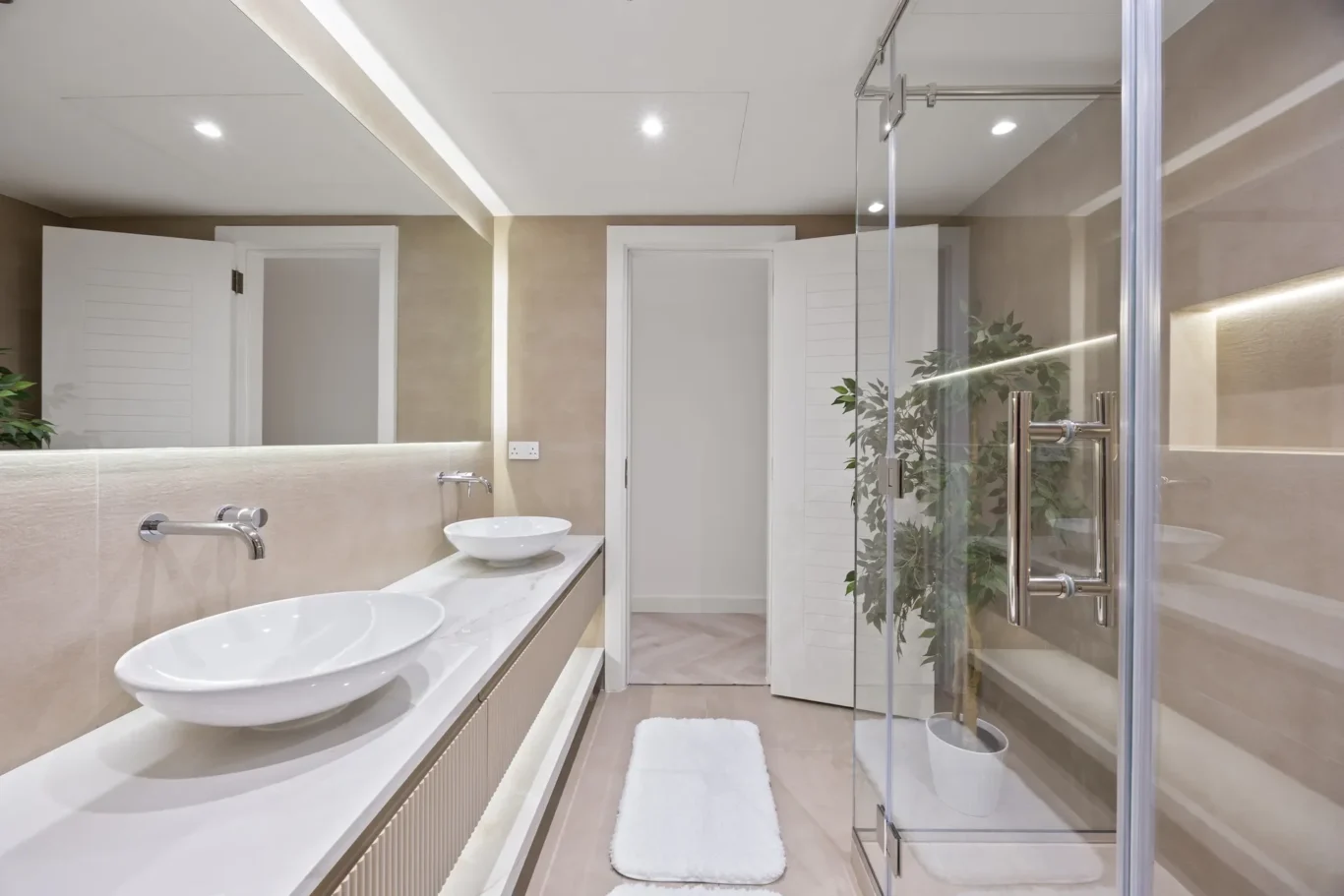 Contemporary bathroom with dual white vessel sinks on a marble counter, beige tiled walls, and a glass shower enclosure.