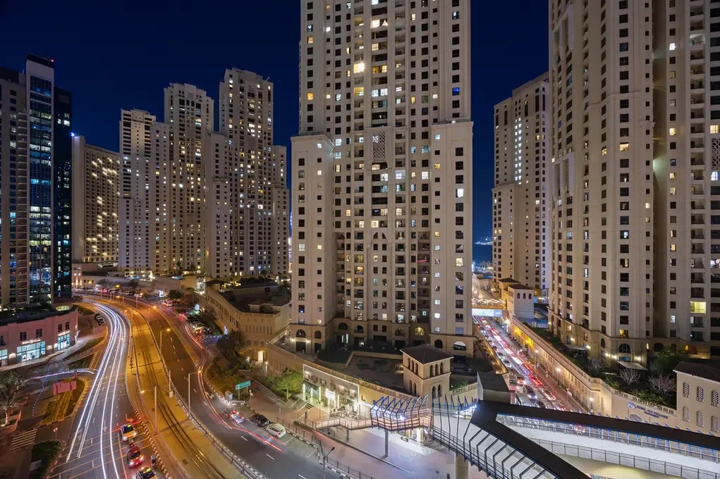 Night cityscape with tall apartment towers and light trails along a curved road below in the evening glow