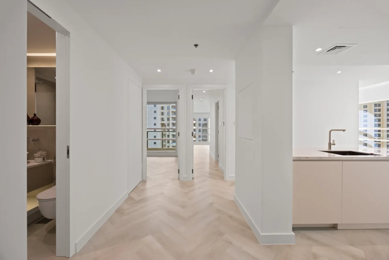 Bright white hallway with light wood chevron floors, leading to glass doors; kitchen area on the right.