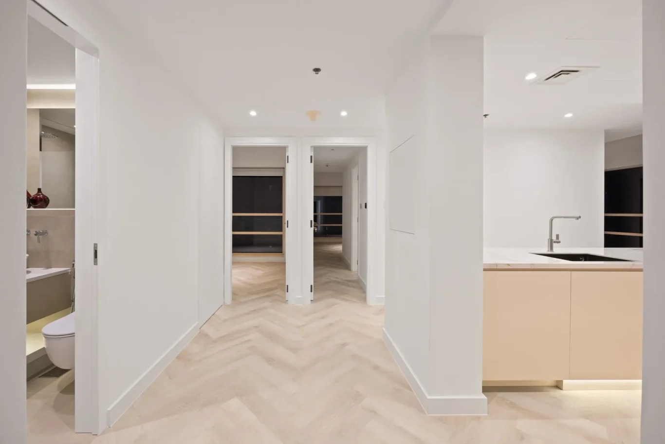 Bright, modern hallway with pale wood flooring leading to doors at the end; open kitchen area on the right with a sink and light cabinetry.