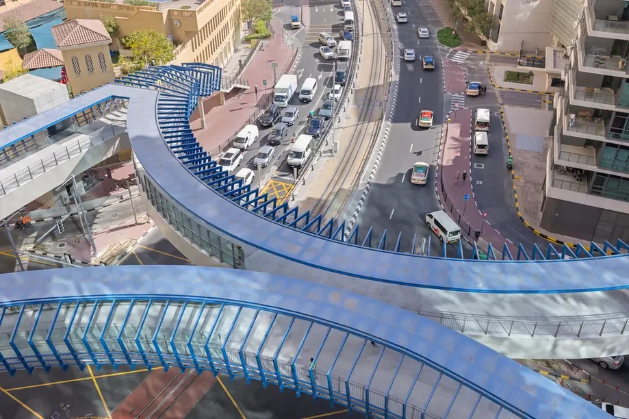 Curved blue elevated pedestrian bridge spanning a busy city street with cars below.