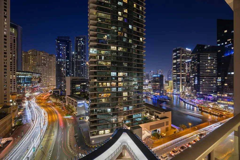 Nighttime cityscape with illuminated skyscrapers along a canal, viewed from a balcony, with light trails from moving cars on the streets.