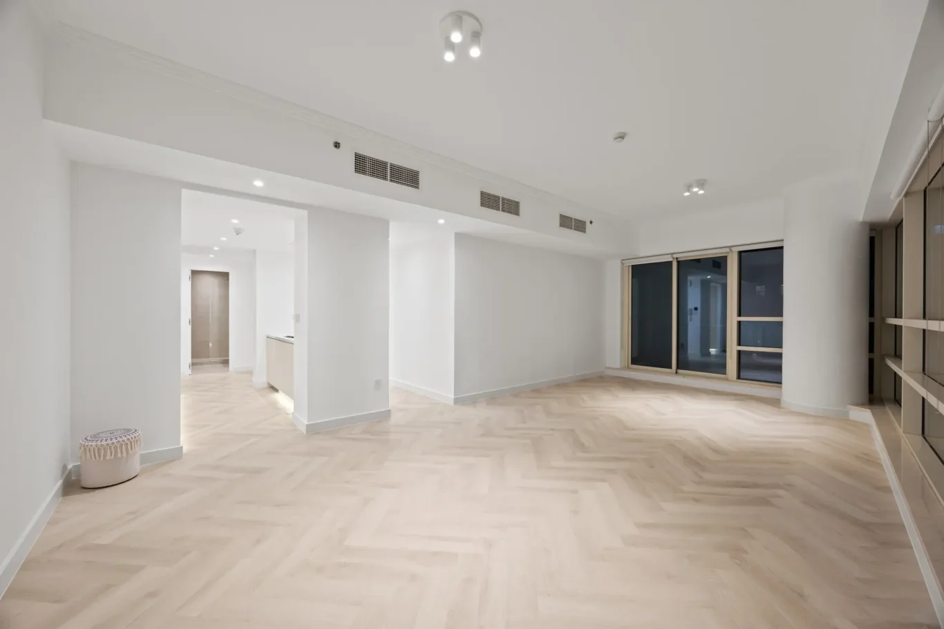 Spacious, minimalist living area with white walls, light wood herringbone floor, and large sliding glass doors to a balcony on the right.