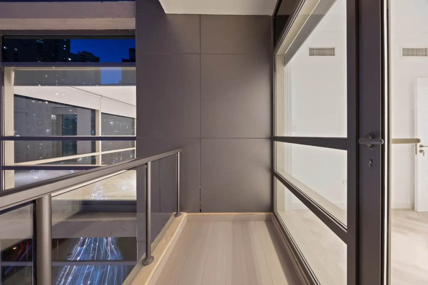 Interior corridor in a modern building at night, featuring glass railing and large windows showing the city lights outside
