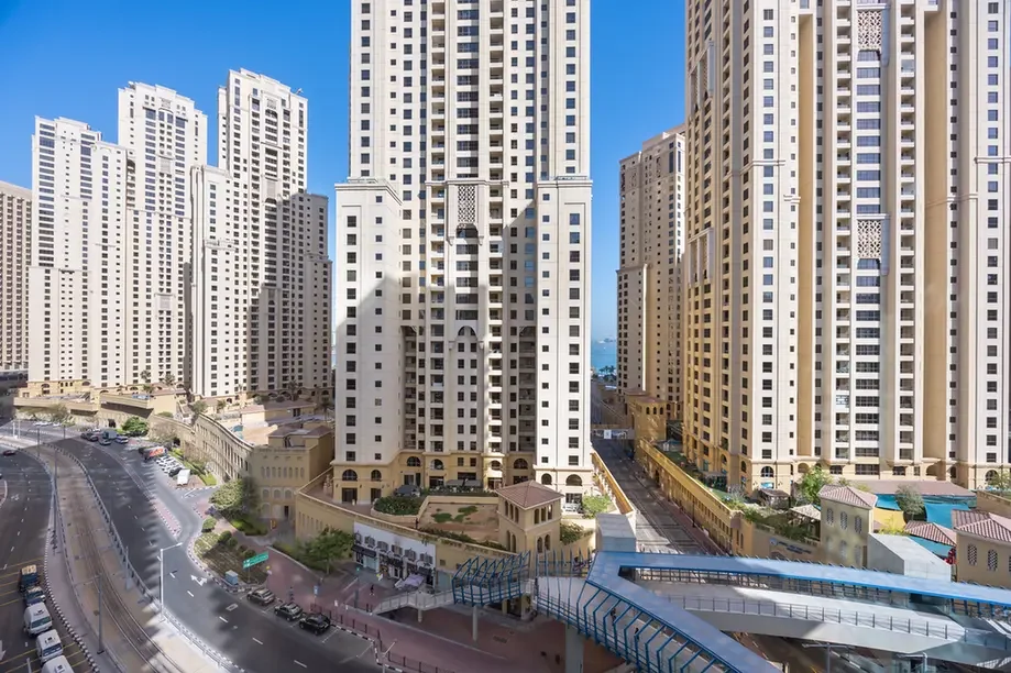 Cluster of tall beige apartment towers lining a curved road under a clear blue sky, with a pedestrian bridge in the foreground.