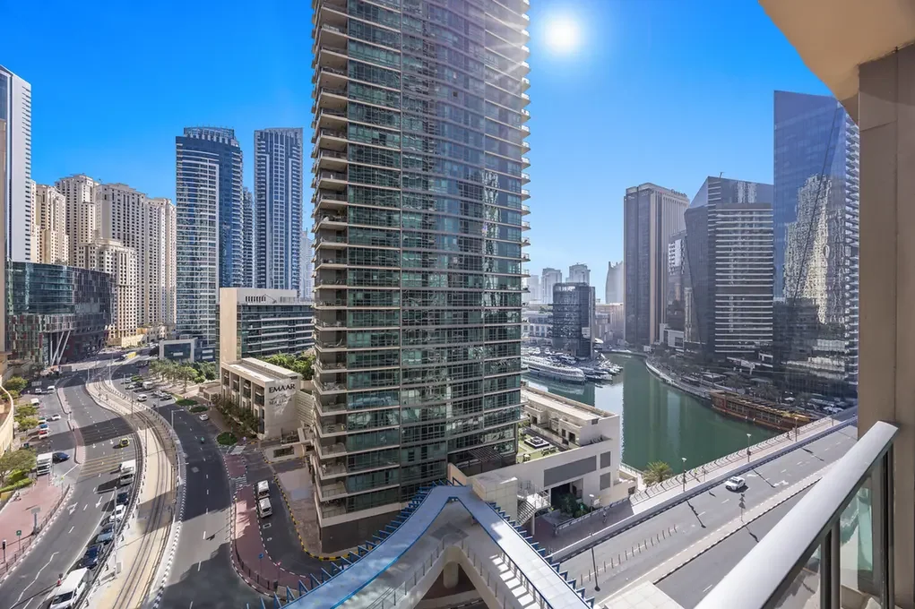 View of a modern city skyline with tall glass buildings, a canal, and roads, seen from a balcony on a sunny day.