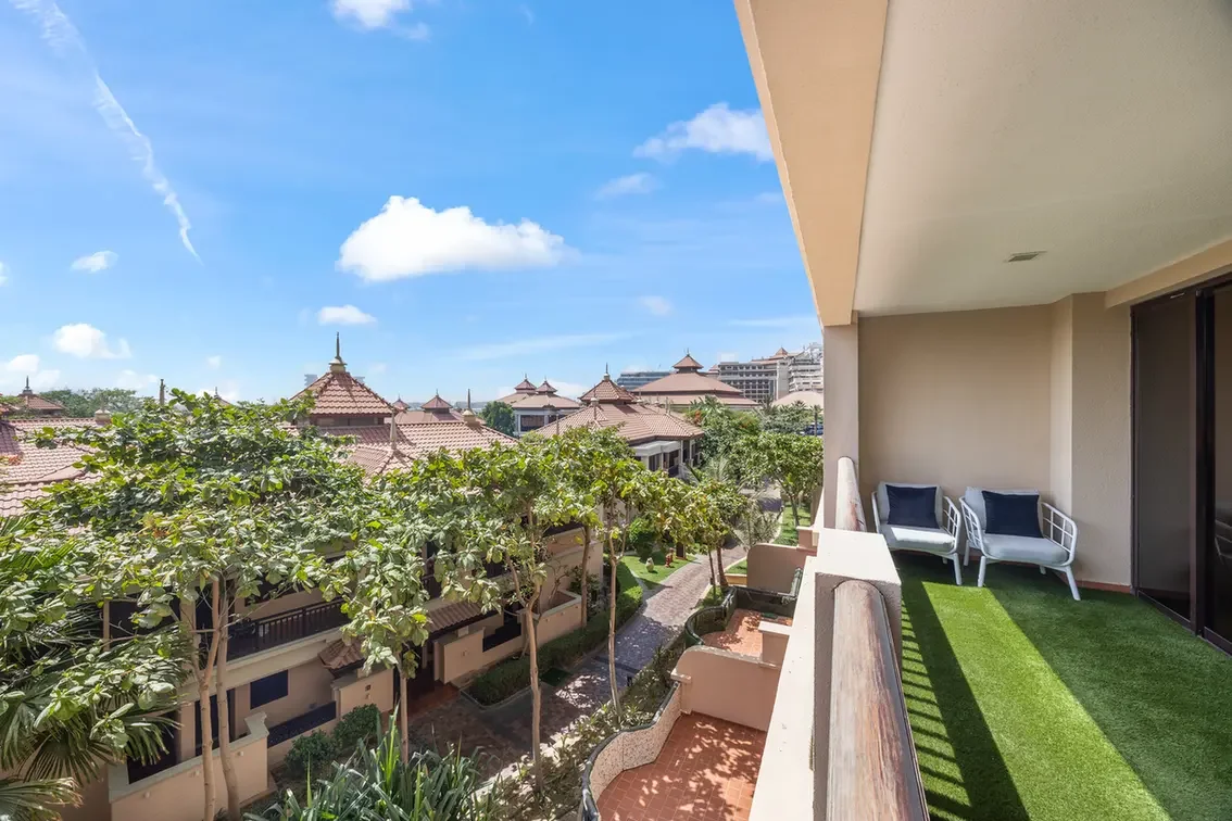 Balcony seating on artificial grass with white chairs, overlooking a courtyard of trees and terracotta-roofed buildings under a blue sky.