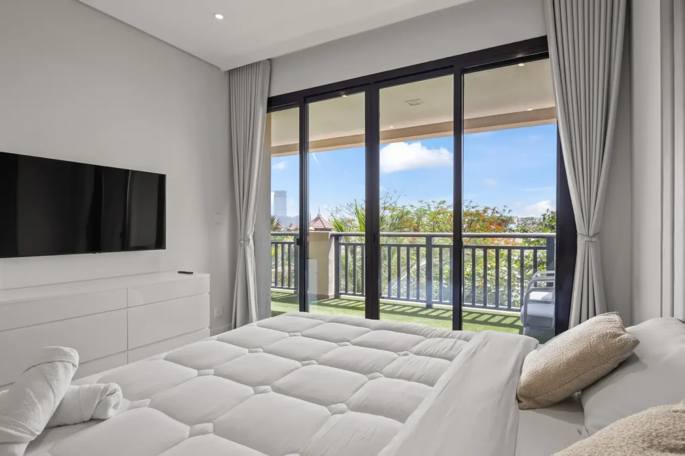 Bright modern bedroom with a large sliding glass door to a balcony, white furnishings, and a wall-mounted TV above a white dresser; view of trees and blue sky outside