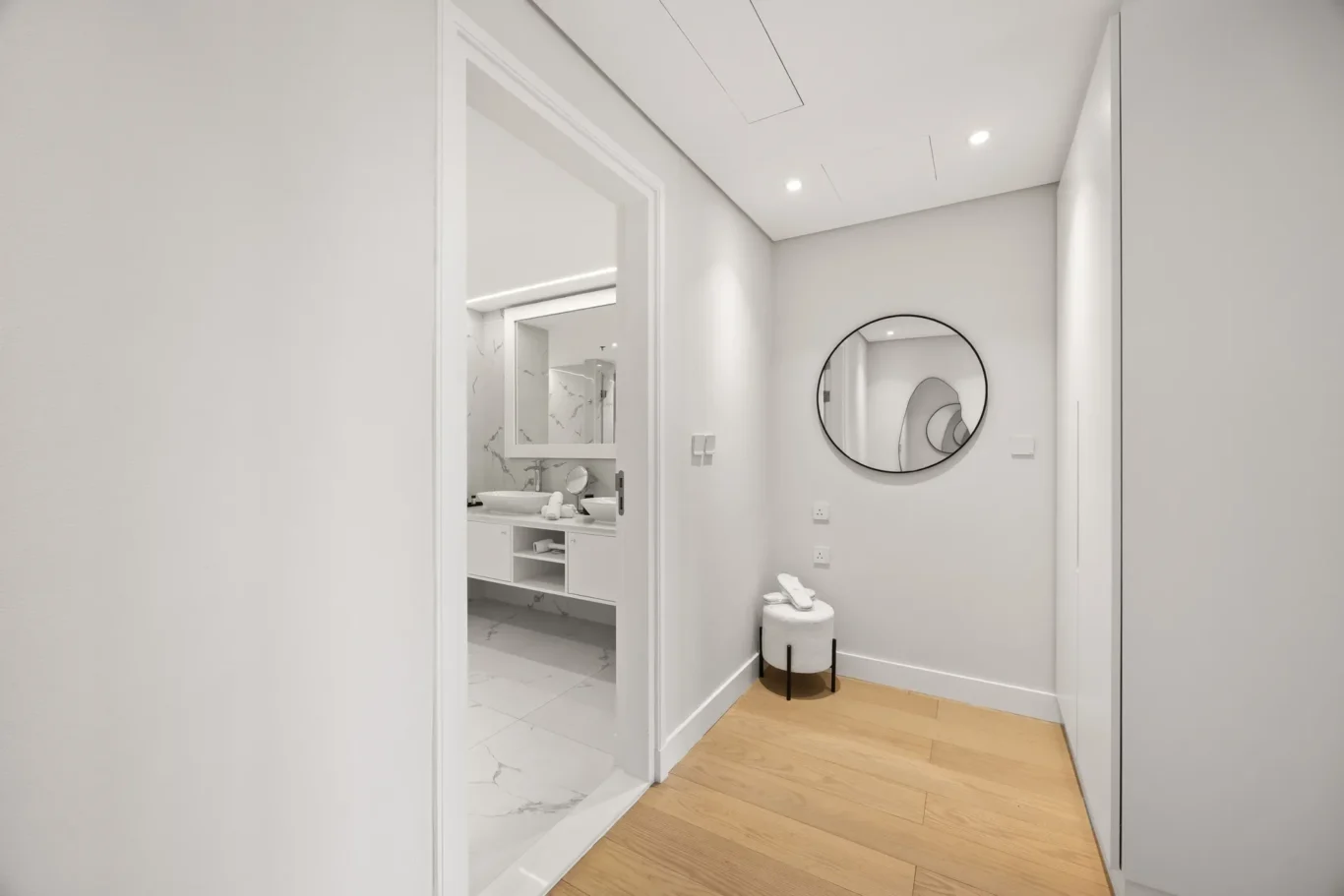 Narrow hallway with light wood floor leading to a white marble bathroom; round black mirror on the wall and a small stool with towels nearby.