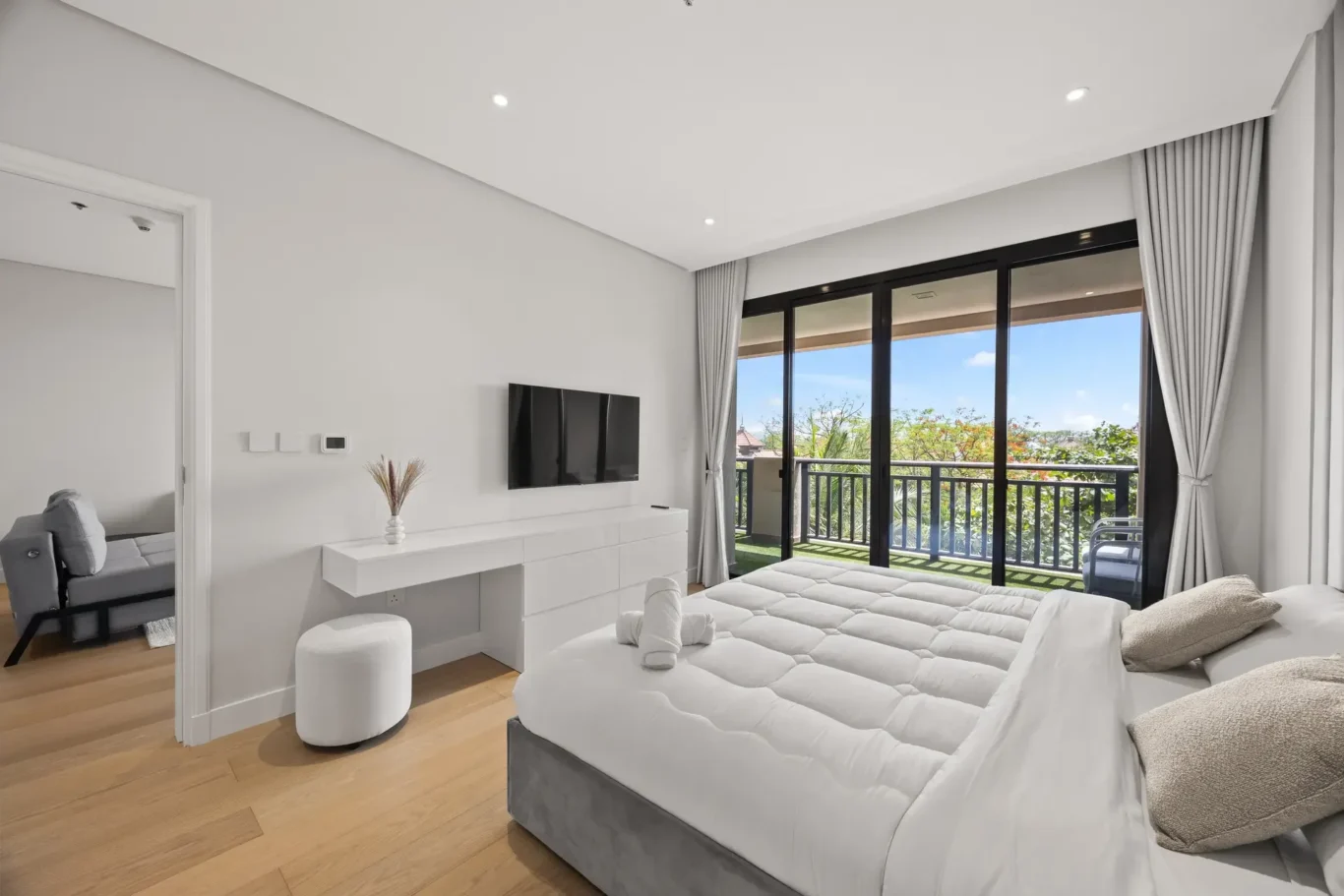 Bright, modern bedroom with white bedding, a wall-mounted TV, and a white dresser, plus large sliding glass doors to a balcony.