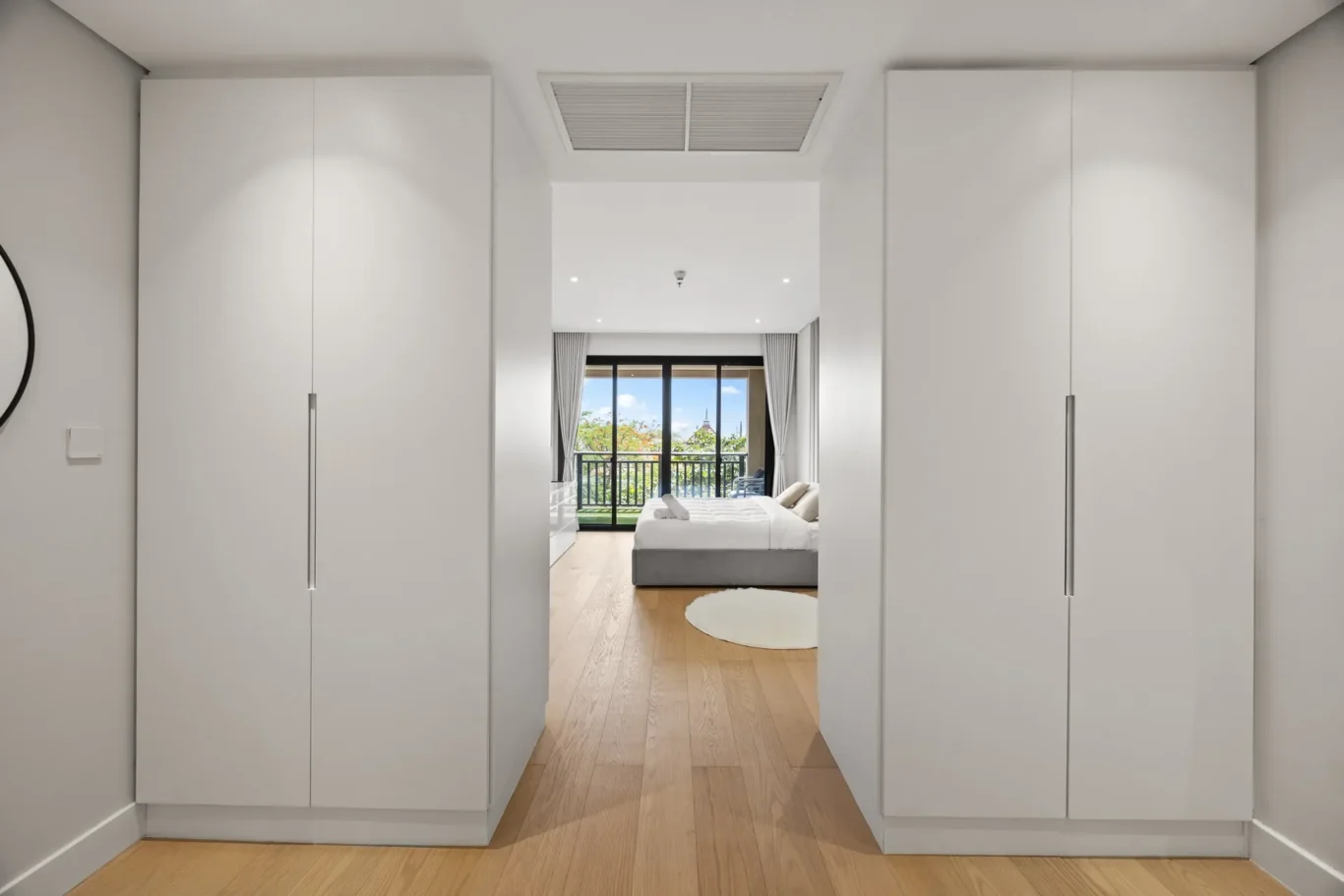 Hallway with white wardrobes on both sides opening to a bright bedroom with a balcony beyond and a wooden floor.