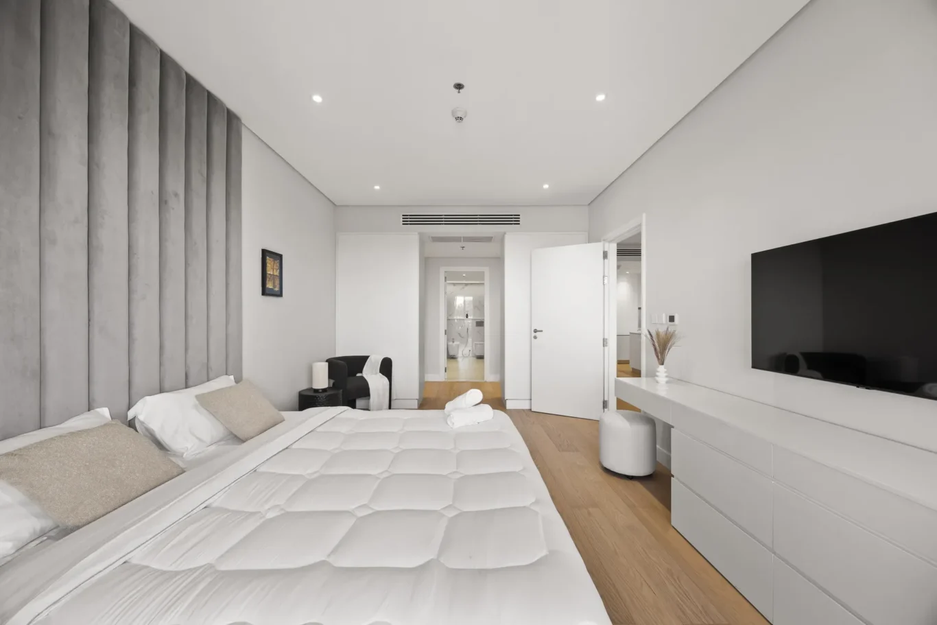 Hotel bedroom with two white beds, a gray padded headboard wall, and a large wall-mounted TV across from the beds.