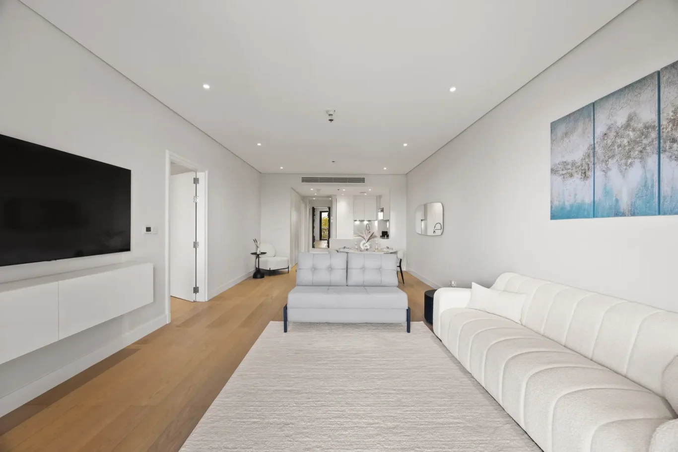 Open-plan living room with light wood floor, white seating, wall-mounted TV on the left, and a blue abstract triptych on the right.
