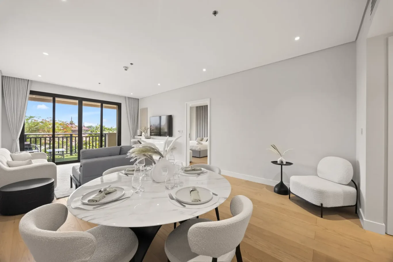 Open-plan living and dining room with a round marble dining table set for six, gray sofas, and sliding glass doors to a balcony.