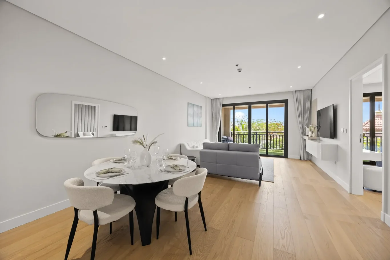 Open-plan living and dining area with a round marble dining table for six, beige chairs, and a gray sofa facing a wall-mounted TV near large glass doors to a balcony.