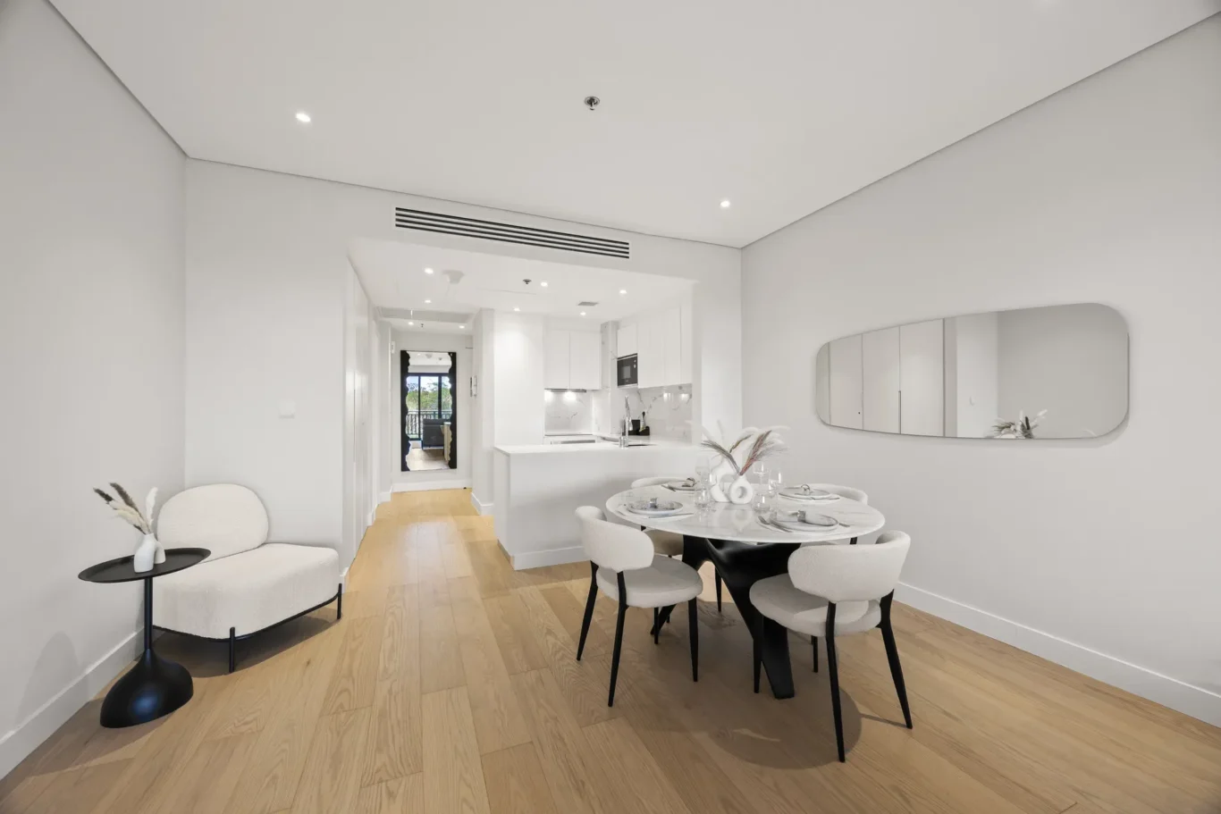 Open-plan dining area with a round table set for six, white kitchen in the background, and a curved wall mirror on the right.