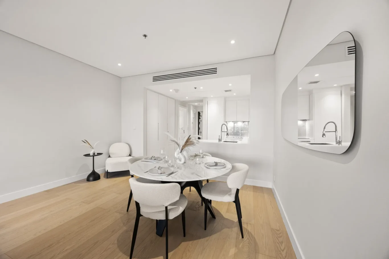 Modern minimalist dining room with a round marble table set for six and light gray upholstered chairs; a curved wall mirror on the right adds depth.