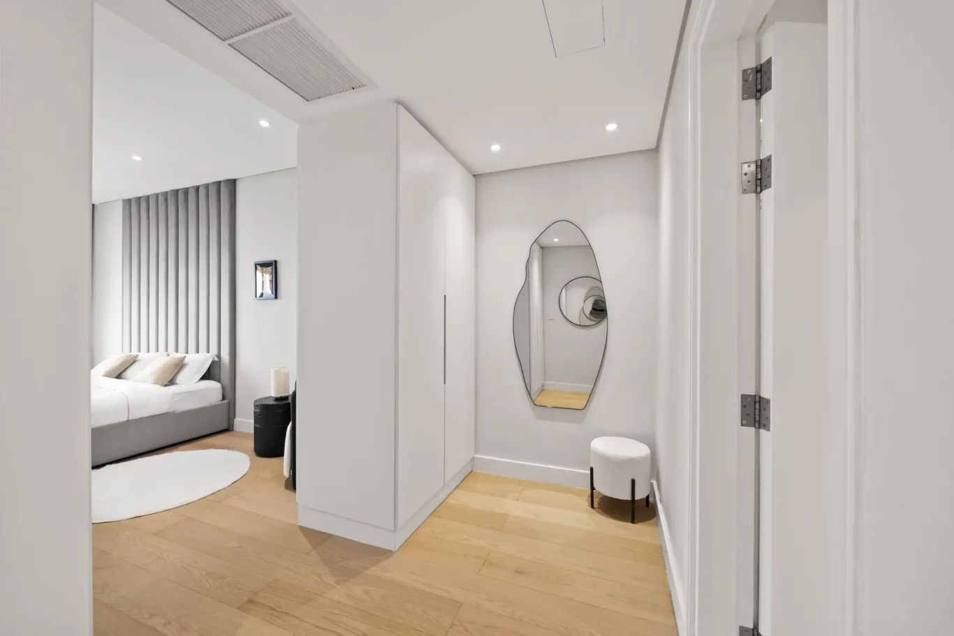 Modern minimalist bedroom with white walls, light wood floors, a tall white wardrobe, and a bed with a grey upholstered headboard visible in the background.