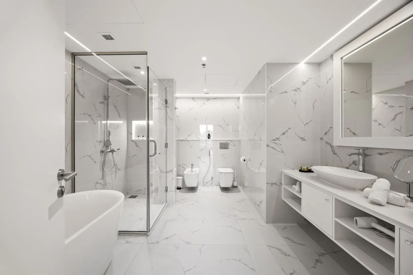 Bright modern bathroom with white marble walls and floor, glass shower enclosure on the left, and a vanity with a vessel sink on the right.