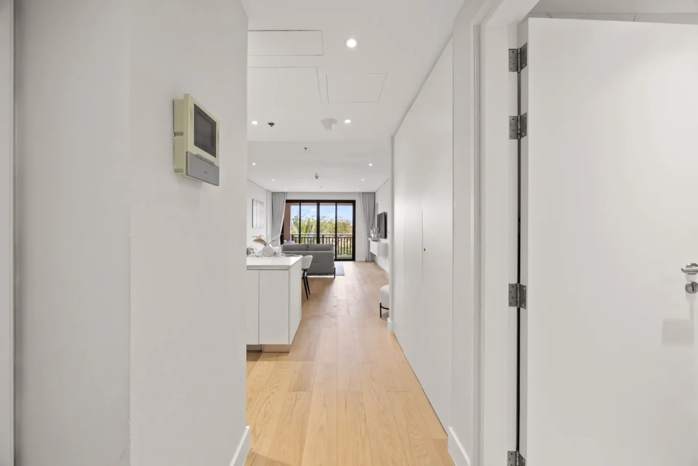 Long white hallway leads to a bright living area with a balcony, open-plan kitchen to the left, and wooden floors.