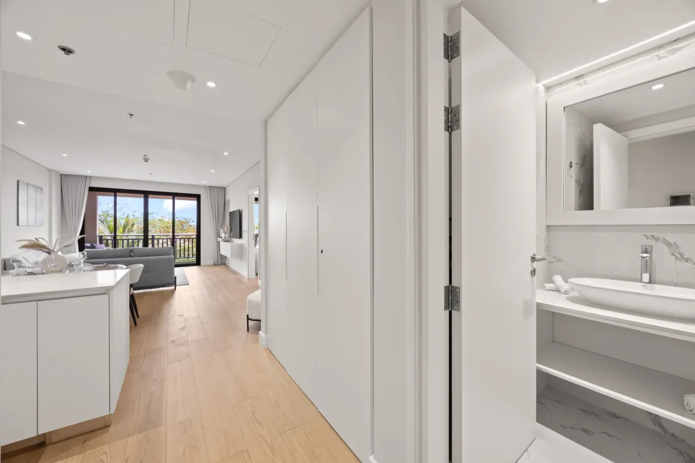 Bright open-plan living area with white cabinets, a gray sofa, and a balcony door open to greenery outside.