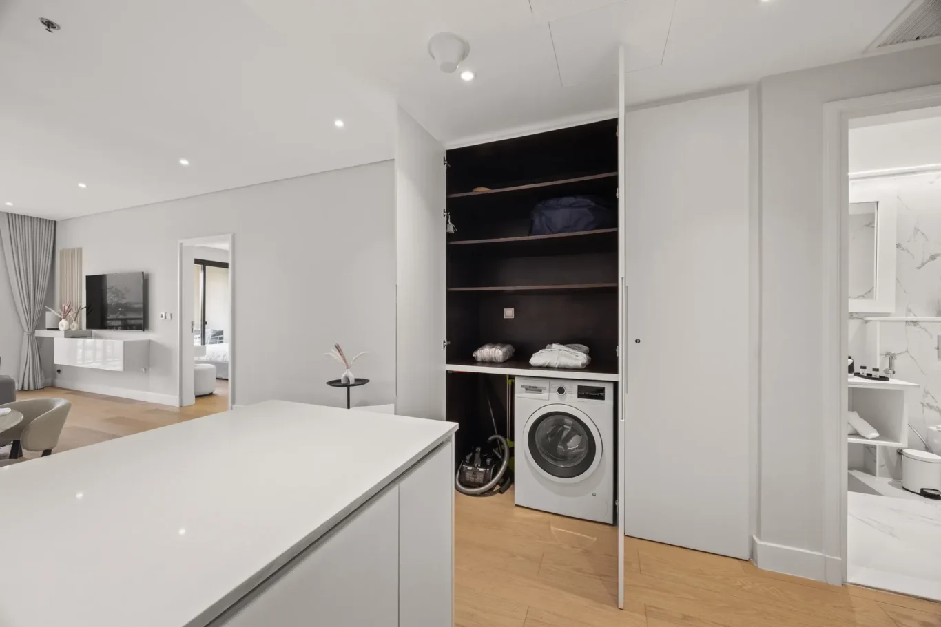 Modern open-plan living area with white kitchen island and a laundry closet housing a front-loading washer and dark shelves above it.