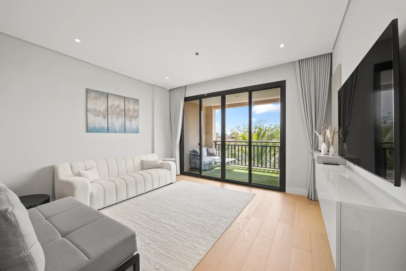 Modern living room with a light gray sectional sofa, abstract triptych art, and large glass sliding doors to a balcony.