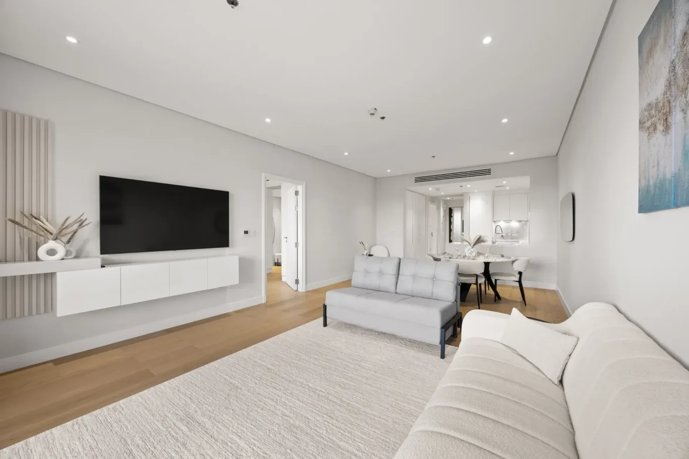 Open-plan living area with a wall-mounted TV, white storage unit, gray sofa, and a dining table near a bright kitchen.