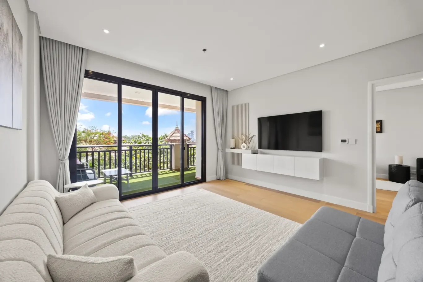Bright modern living room with a wall-mounted flat-screen TV, white floating cabinet, and light gray sofa facing large sliding glass doors to a balcony.