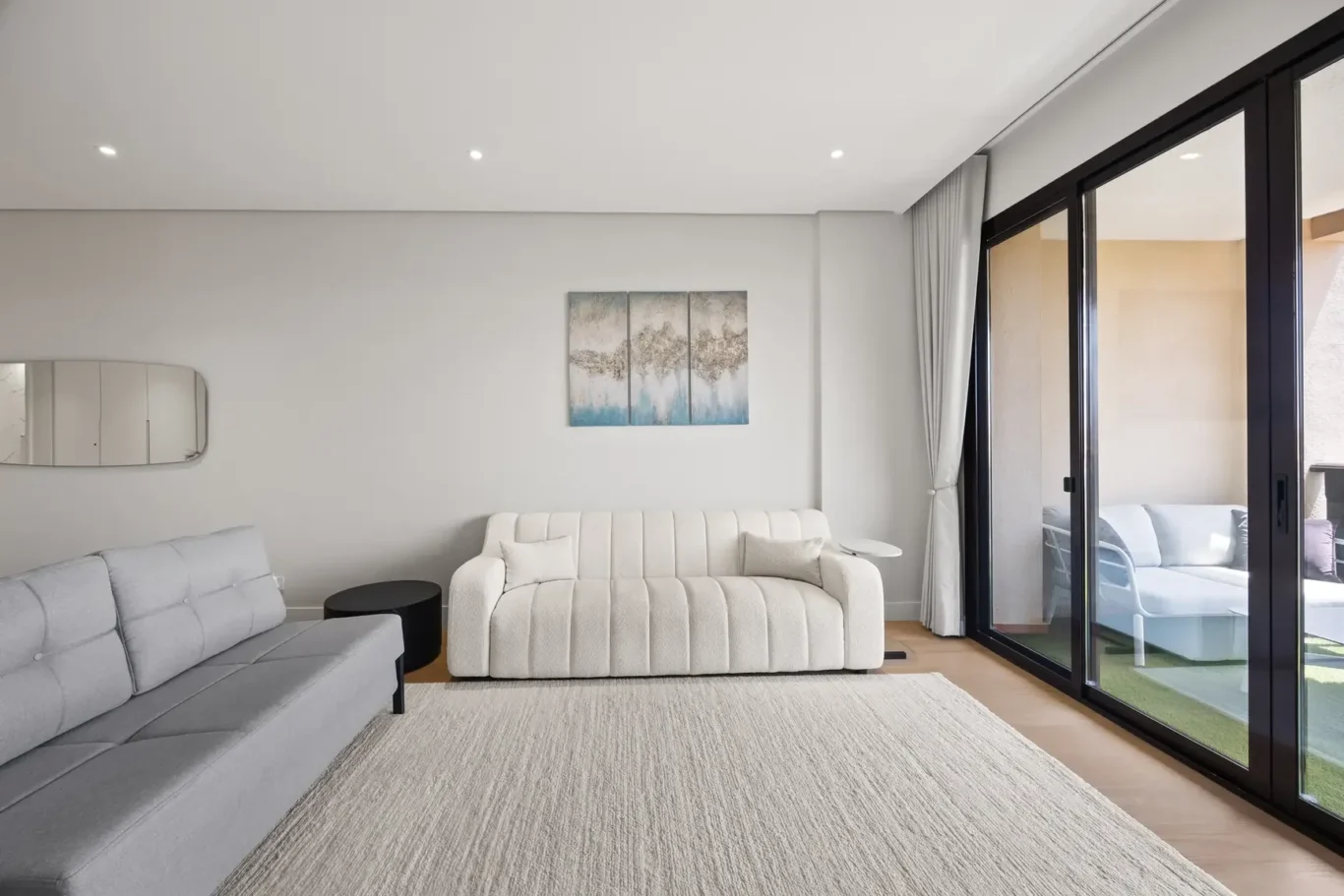 Bright minimalist living room with light gray sofas, a three-panel abstract mural, and floor-to-ceiling glass doors opening to an outdoor seating area.