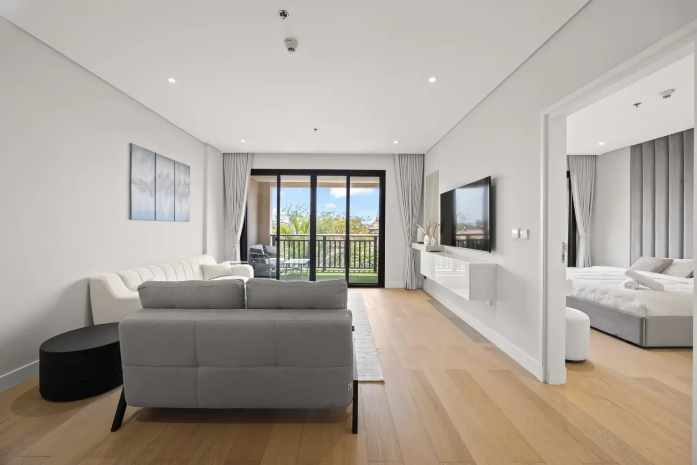 Bright modern living room with a gray sectional sofa, white walls, and a balcony through sliding glass doors with outdoor seating visible outside.