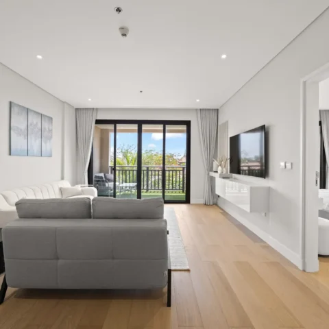 Bright modern living room with a gray sectional sofa, white walls, and a balcony through sliding glass doors with outdoor seating visible outside.