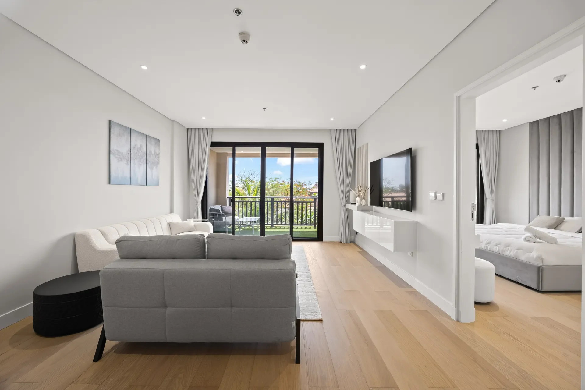 Bright modern living room with a gray sectional sofa, white walls, and a balcony through sliding glass doors with outdoor seating visible outside.
