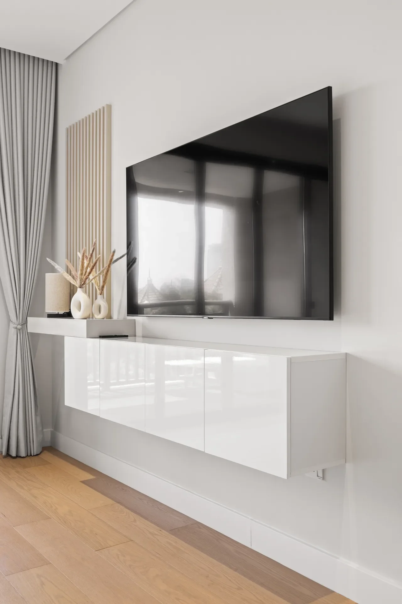 Wall-mounted flat-screen TV above a glossy white storage cabinet with decorative vases and dried grasses; gray curtains on the left.