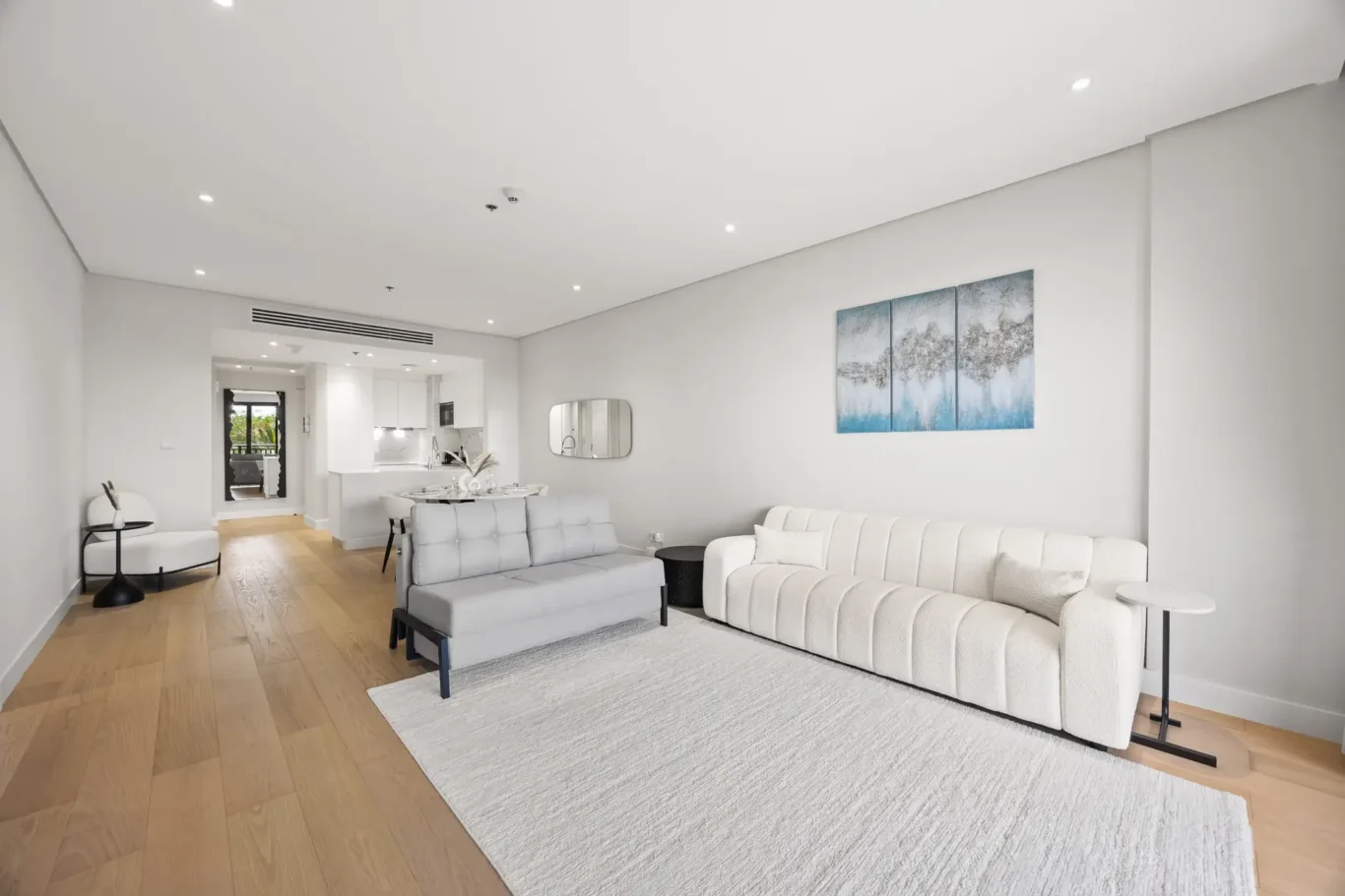 Open-concept living space with a gray sofa, white tufted sofa, light wood floors, and a blue-gray 3-panel wall art above a white wallMirror visible in the background near the kitchen.