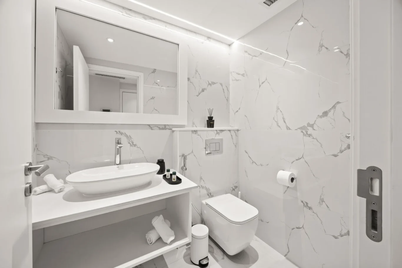 White minimalist bathroom with marble walls, a vessel sink on a floating vanity, and a large rectangular mirror.