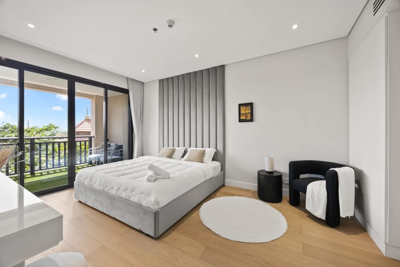 Bright hotel bedroom with a king bed, gray upholstered headboard wall, and balcony doors with outdoor seating visible outside.