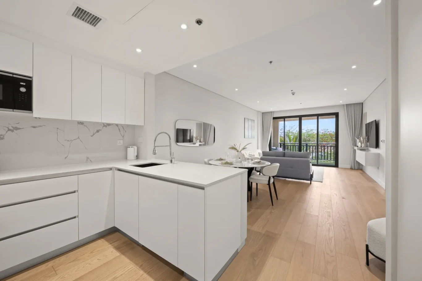 Bright open-plan kitchen and living area with white cabinets, marble backsplash, and a balcony view through large doors.
