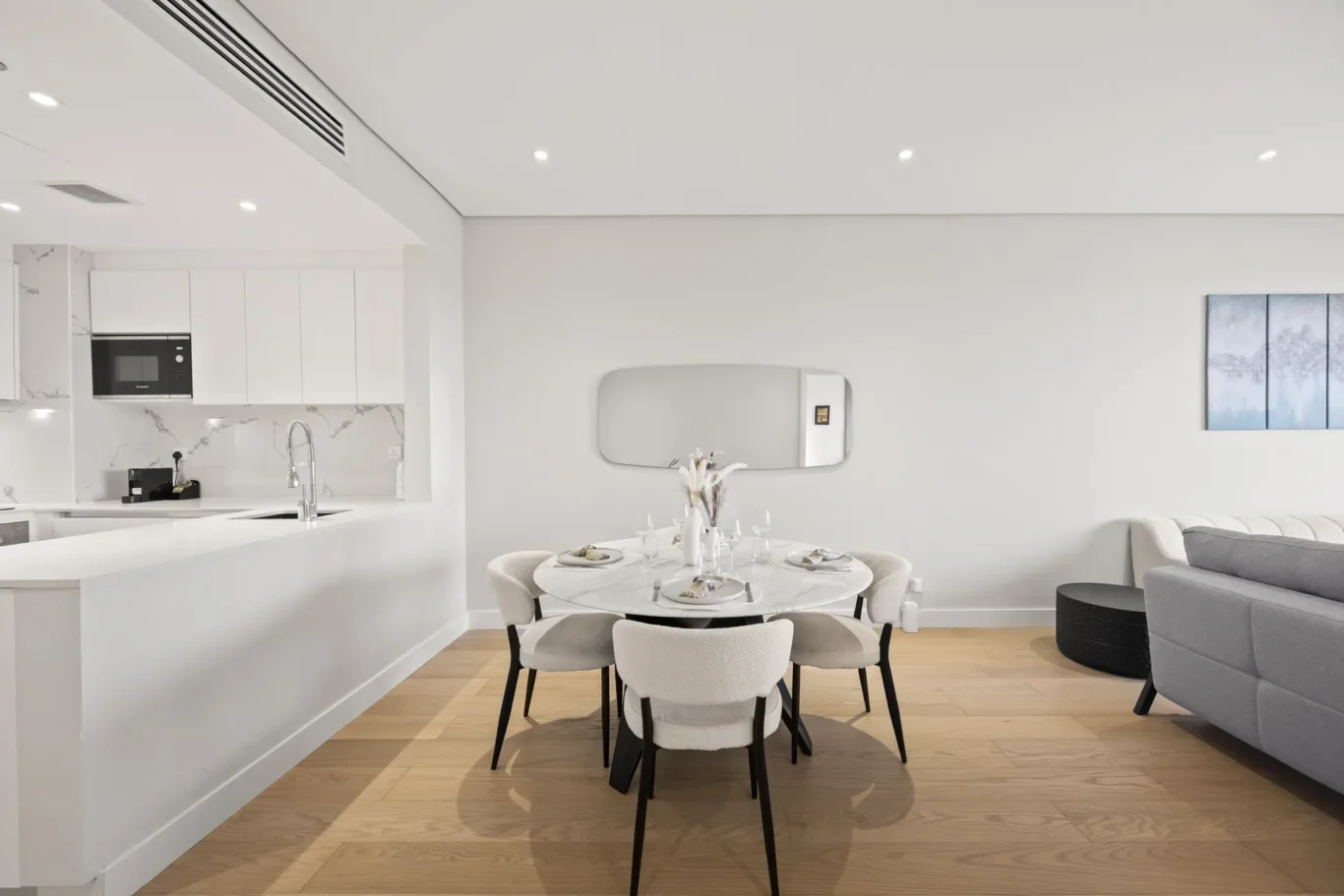 Open-plan kitchen and dining area with white cabinetry, marble accents, and a round table set for six in a bright living space.